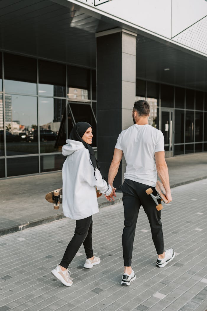 A young couple holding hands and skateboards, walking outdoors in a modern urban area.